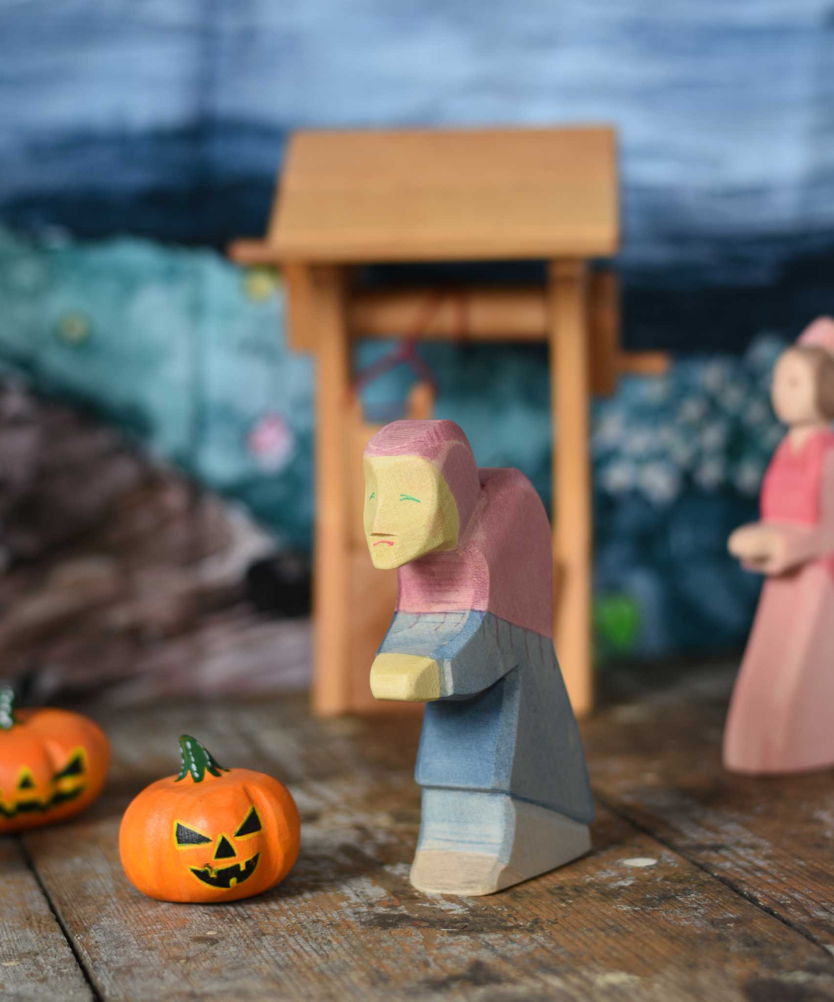 An Ostheimer hand made and hand painted wooden witch figure placed on a wooden surface in a small world play scene. An Ostheimer wooden well and other wooden toys including Bumbu pumpkins can be seen in the background. These wooden figures are part of a wide range of expertly hand crafted wooden toys here at Babipur.