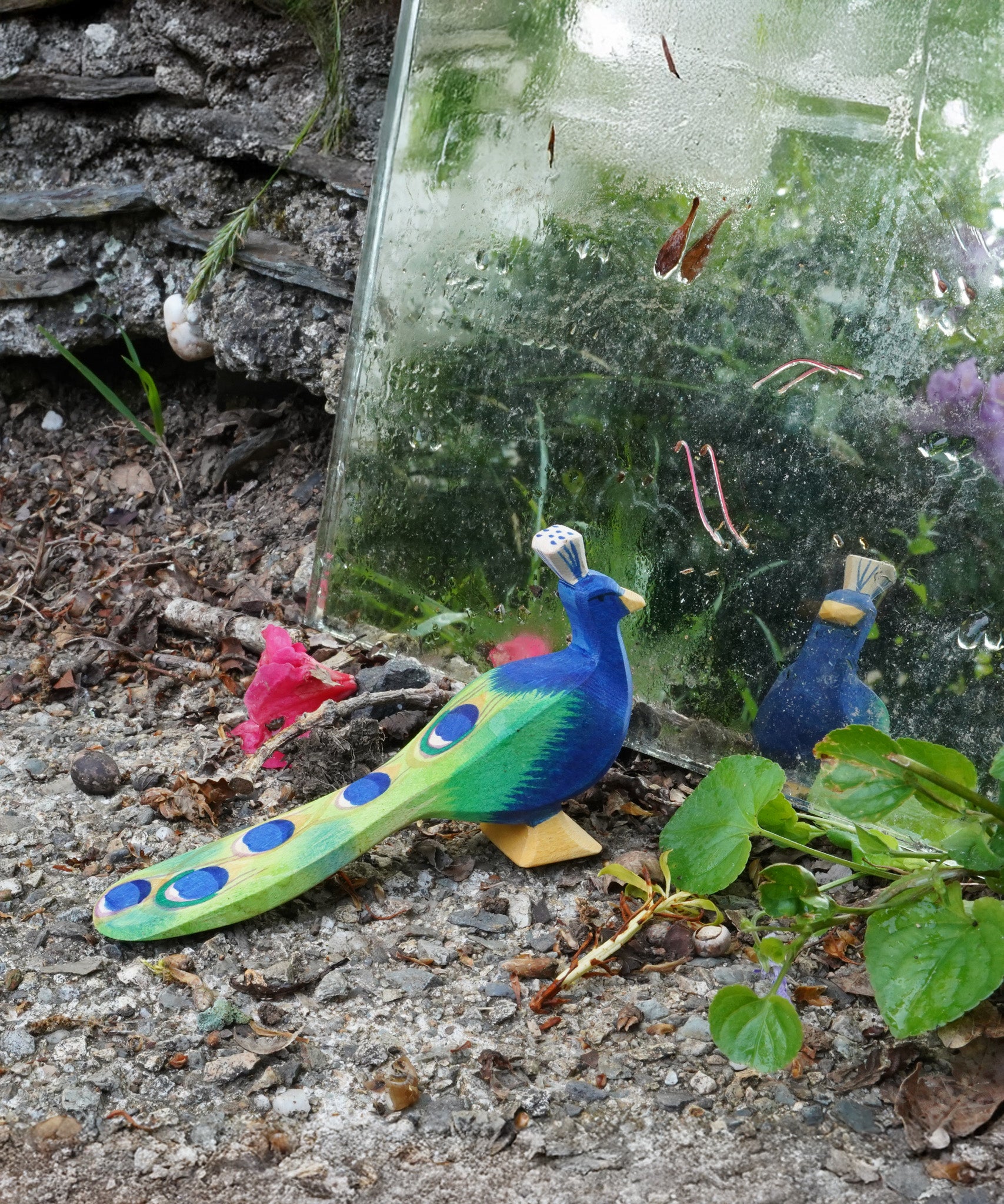 Ostheimer handmade wooden peacock figure with it's feathers closed standing in front of glass. Figure is hand-painted in vibrant blues and greens, made from sustainable wood in Germany and sold at Babipur.