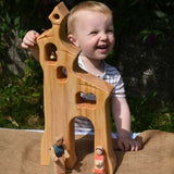 A child smiling standing behind an Ostheimer wooden bell tower. The child is reaching up for the bell. These wooden figures are part of a large range of expertly hand crafted, Waldorf inspired, wooden toys here at Babipur.