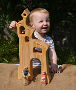 A child smiling standing behind an Ostheimer wooden bell tower. The child is reaching up for the bell. These wooden figures are part of a large range of expertly hand crafted, Waldorf inspired, wooden toys here at Babipur.