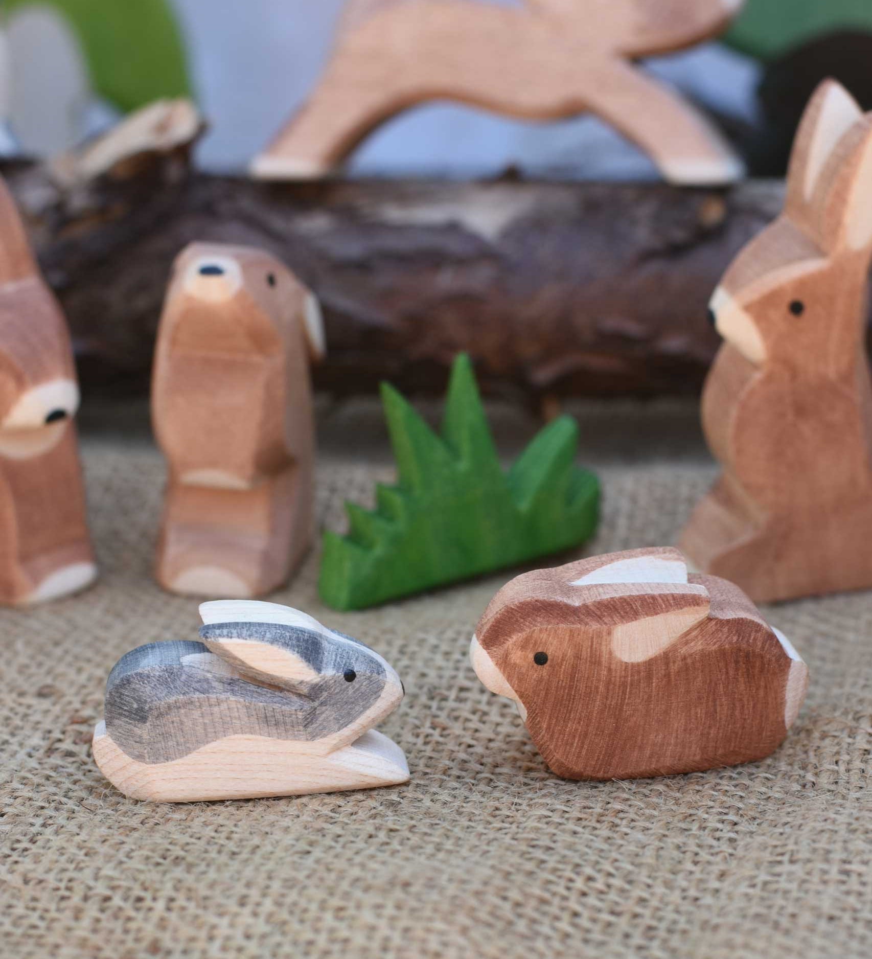 Two small Ostheimer rabbit wooden toy figures placed at the front of a small world play scene. One rabbit is  Black and White and the other is brown. Other rabbits from the collection can be seen in the background. Check out our full range of expertly crafted wooden toy figures and scenery here at Babipur.