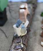 An Ostheimer Wooden Black and White Sitting Rabbit Figure placed on a wooden branch in the middle of a small world play scene. Other Ostheimer toy figures can be seen in the background. 