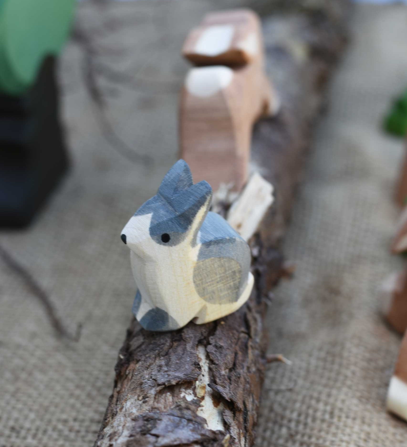 An Ostheimer Wooden Black and White Sitting Rabbit Figure placed on a wooden branch in the middle of a small world play scene. Other Ostheimer toy figures can be seen in the background. 