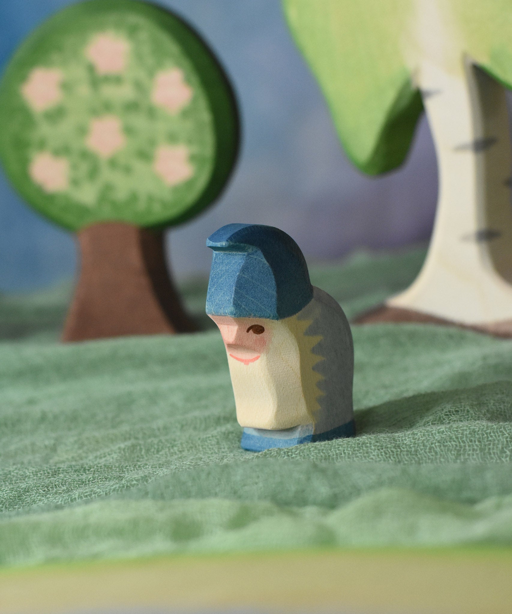 An Ostheimer blue dwarf wooden toy figure placed in a small world play scene. The dwarf figure has a purple coloured outfit with matching hat and a long white beard. The figure has been placed on a green coloured Ostheimer play scarf. This wooden figure is part of a large range of expertly hand crafted, Waldorf inspired, wooden toys here at Babipur. 