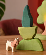 Ostheimer light, middle and dark green wooden bush scenery pieces stacked on top of each other with a small goat figure placed besides them.