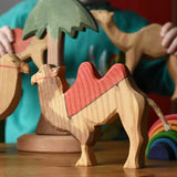 Ostheimer wooden camel figure with red saddle placed on a wooden surface with a tall palm tree in the background and a child playing with other camel figures.