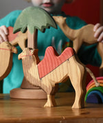 Ostheimer wooden camel figure with red saddle placed on a wooden surface with a tall palm tree in the background and a child playing with other camel figures.