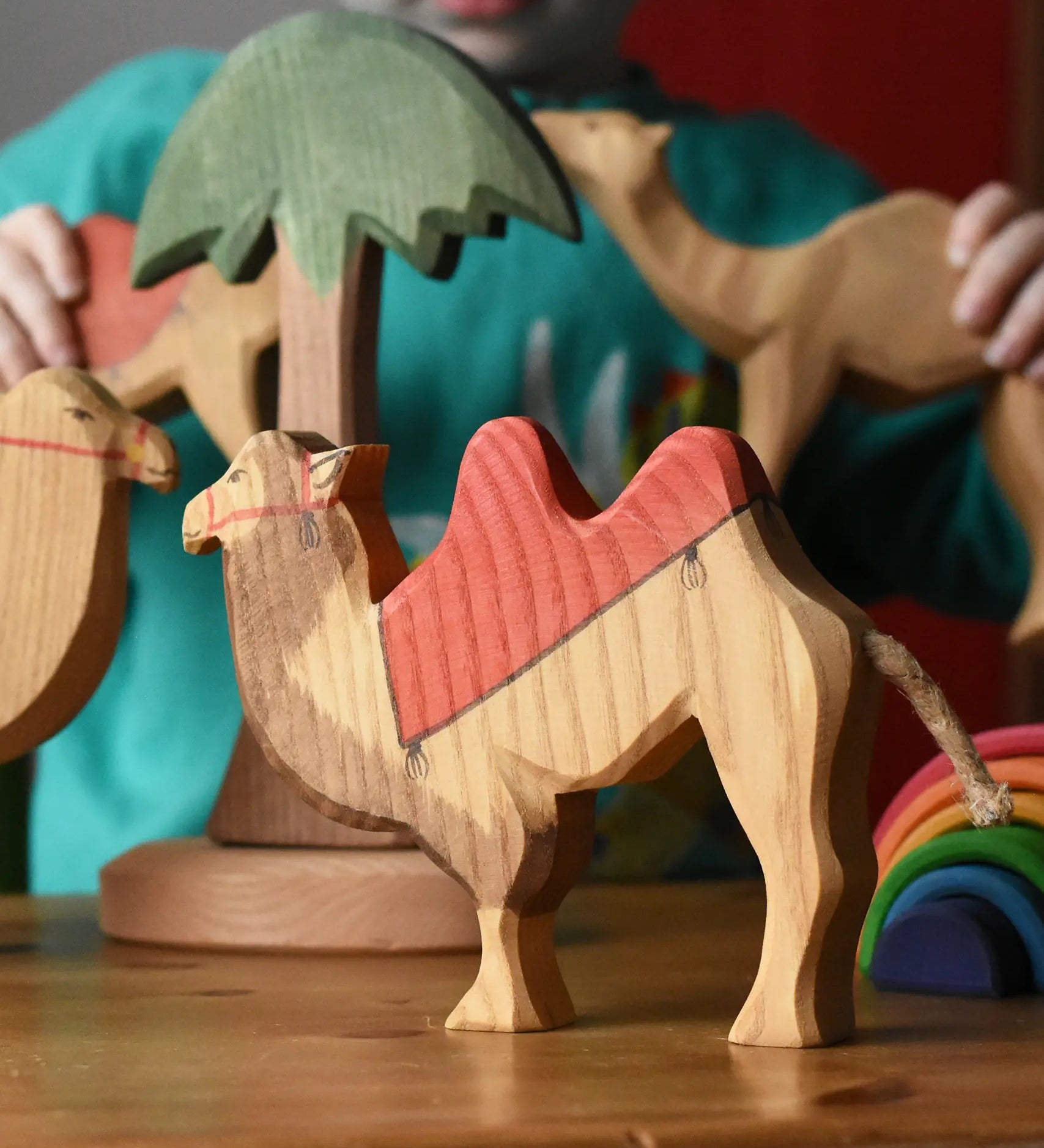 Ostheimer wooden camel figure with red saddle placed on a wooden surface with a tall palm tree in the background and a child playing with other camel figures.