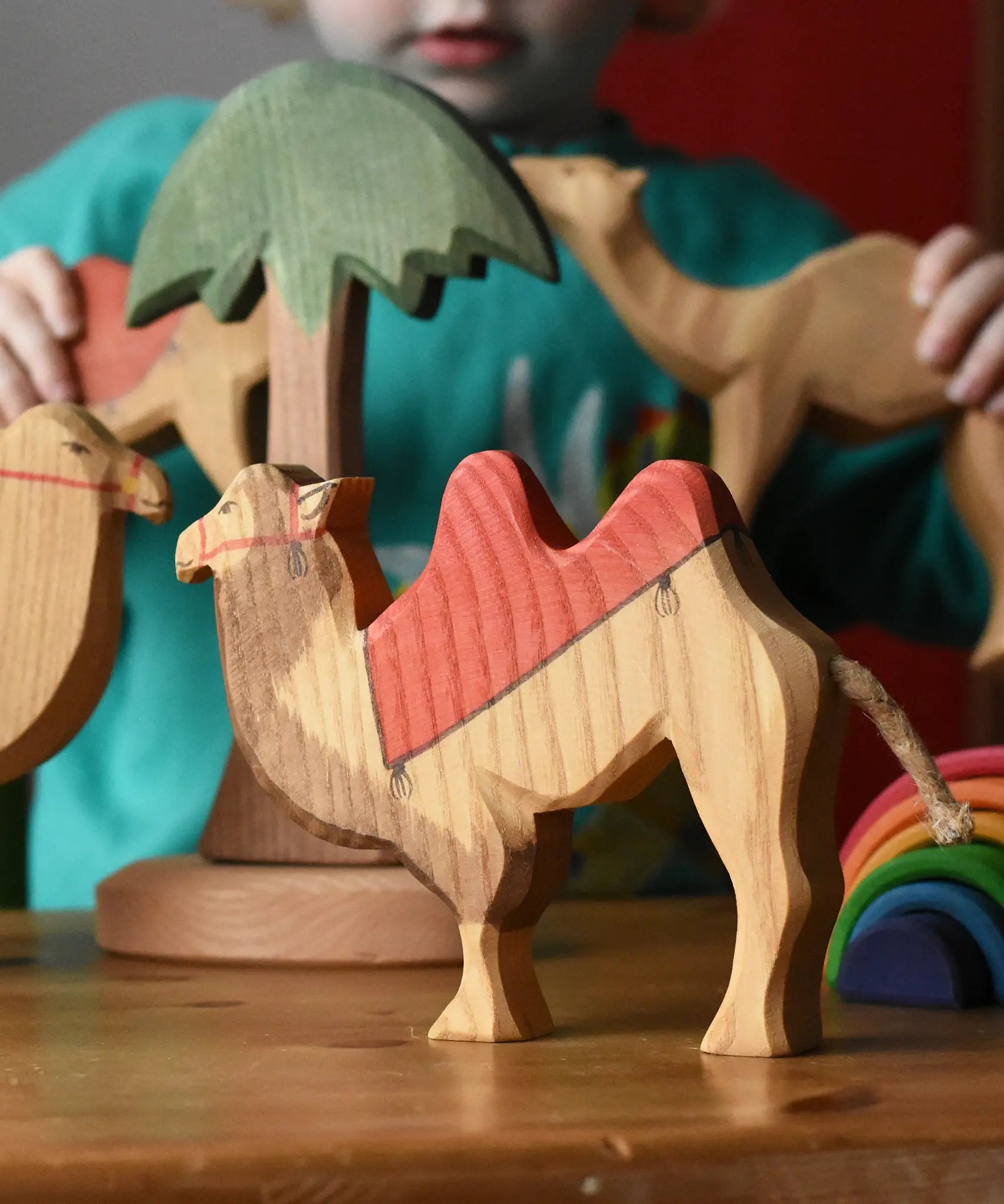 Ostheimer wooden camel figure with red saddle placed on a wooden surface with a tall palm tree in the background and a child playing with other camel figures.