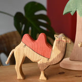 Ostheimer wooden camel figure with red saddle placed on a wooden surface with a tall palm tree in the background.