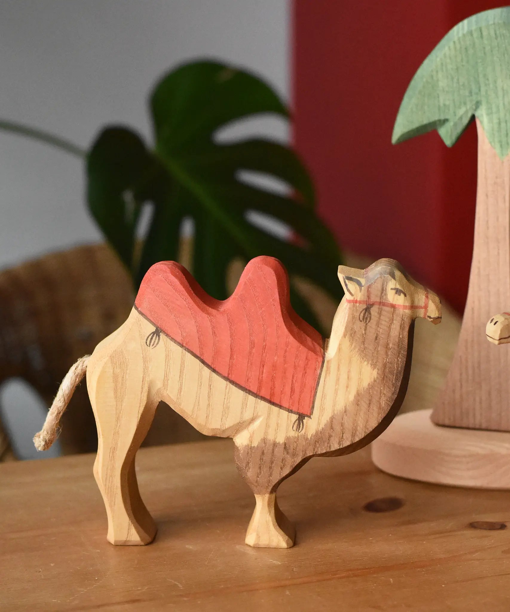 Ostheimer wooden camel figure with red saddle placed on a wooden surface with a tall palm tree in the background.