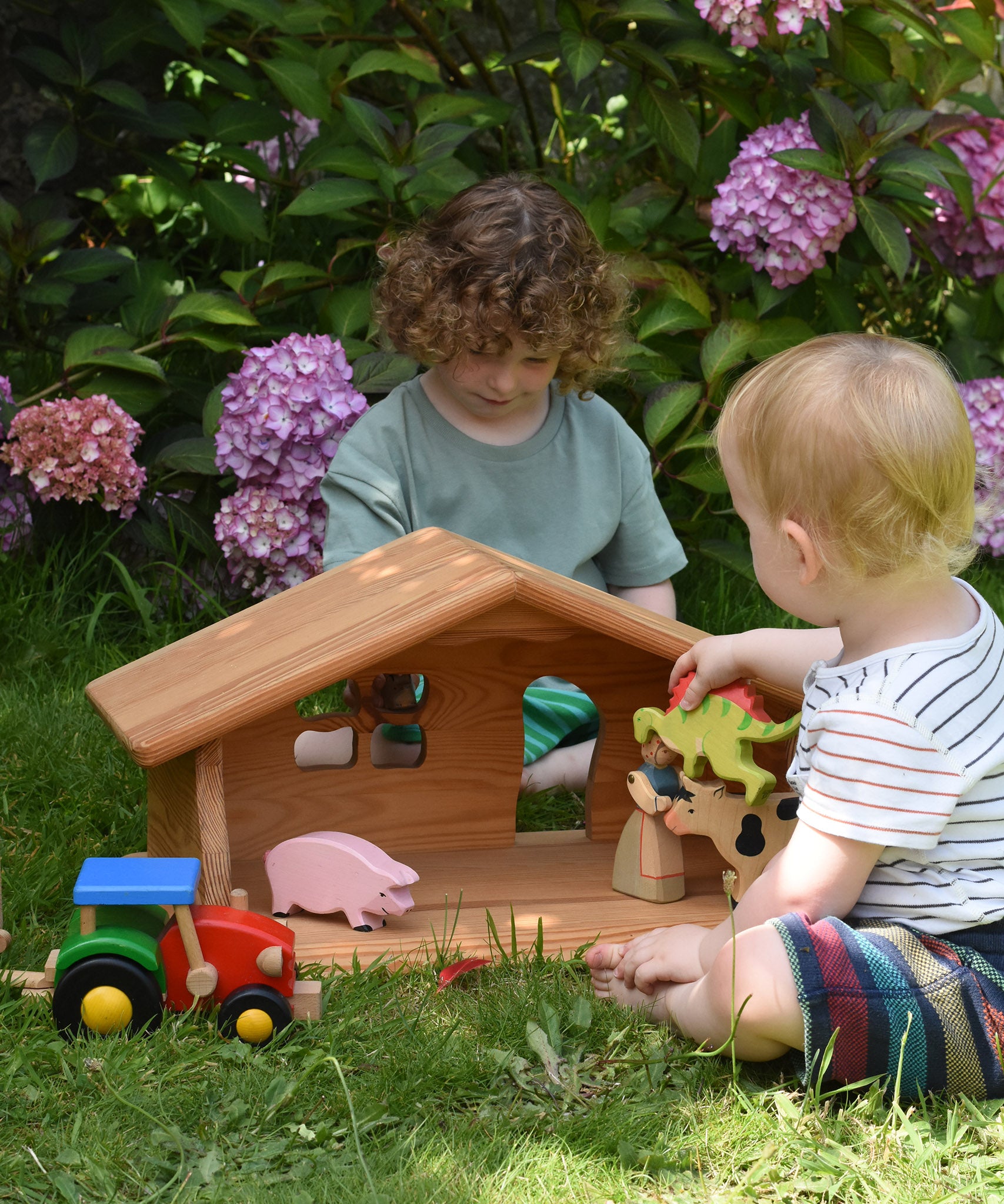 Children sitting on grass outdoors playing with the Ostheimer wooden dolls house. They have wooden animal figures from Holztiger and a wooden tractor by Bajo. These wooden figures are part of a large range of expertly hand crafted, Waldorf inspired, wooden toys here at Babipur.