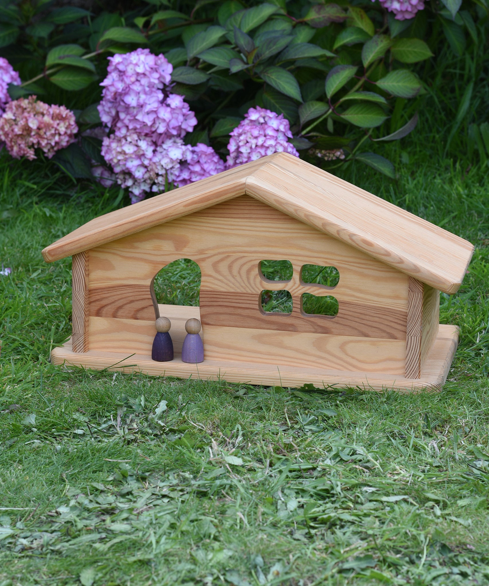 An Ostheimer wooden dolls house placed on grass with two purple Nins stood in the doorway. These wooden figures are part of a large range of expertly hand crafted, Waldorf inspired, wooden toys here at Babipur. 