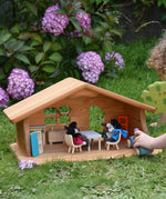 A Ostheimer wooden dolls house set up with PlanToys dolls and furniture inside. A child's hand can be seen to the side driving a Grimm's car around. These wooden figures are part of a large range of expertly hand crafted, Waldorf inspired, wooden toys here at Babipur.