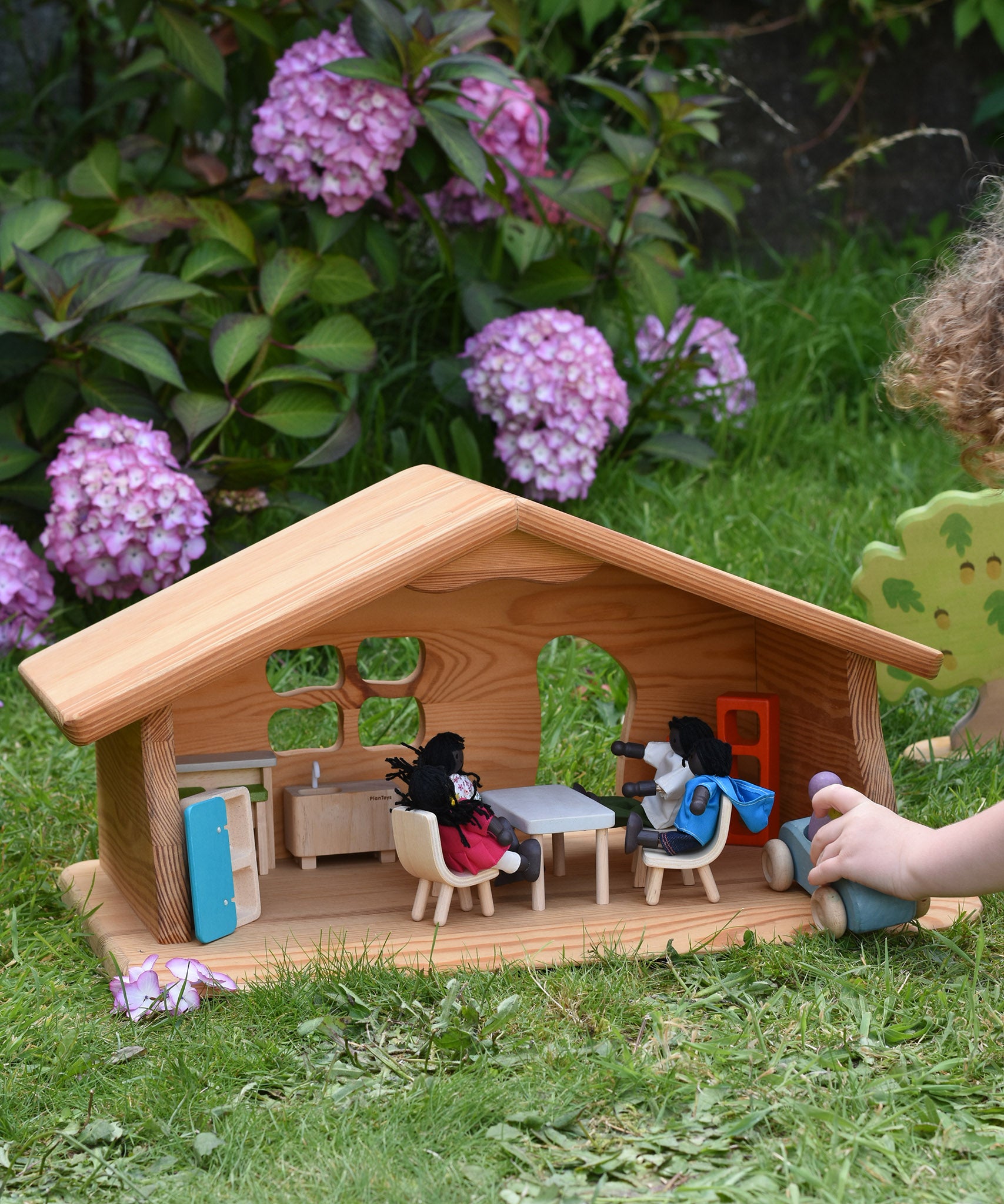 A Ostheimer wooden dolls house set up with PlanToys dolls and furniture inside. A child's hand can be seen to the side driving a Grimm's car around. These wooden figures are part of a large range of expertly hand crafted, Waldorf inspired, wooden toys here at Babipur.