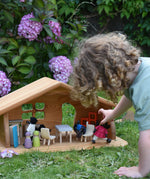 A Ostheimer wooden dolls house set up with PlanToys dolls and furniture inside. A child's hand can be seen holding one of the dolls. These wooden figures are part of a large range of expertly hand crafted, Waldorf inspired, wooden toys here at Babipur.