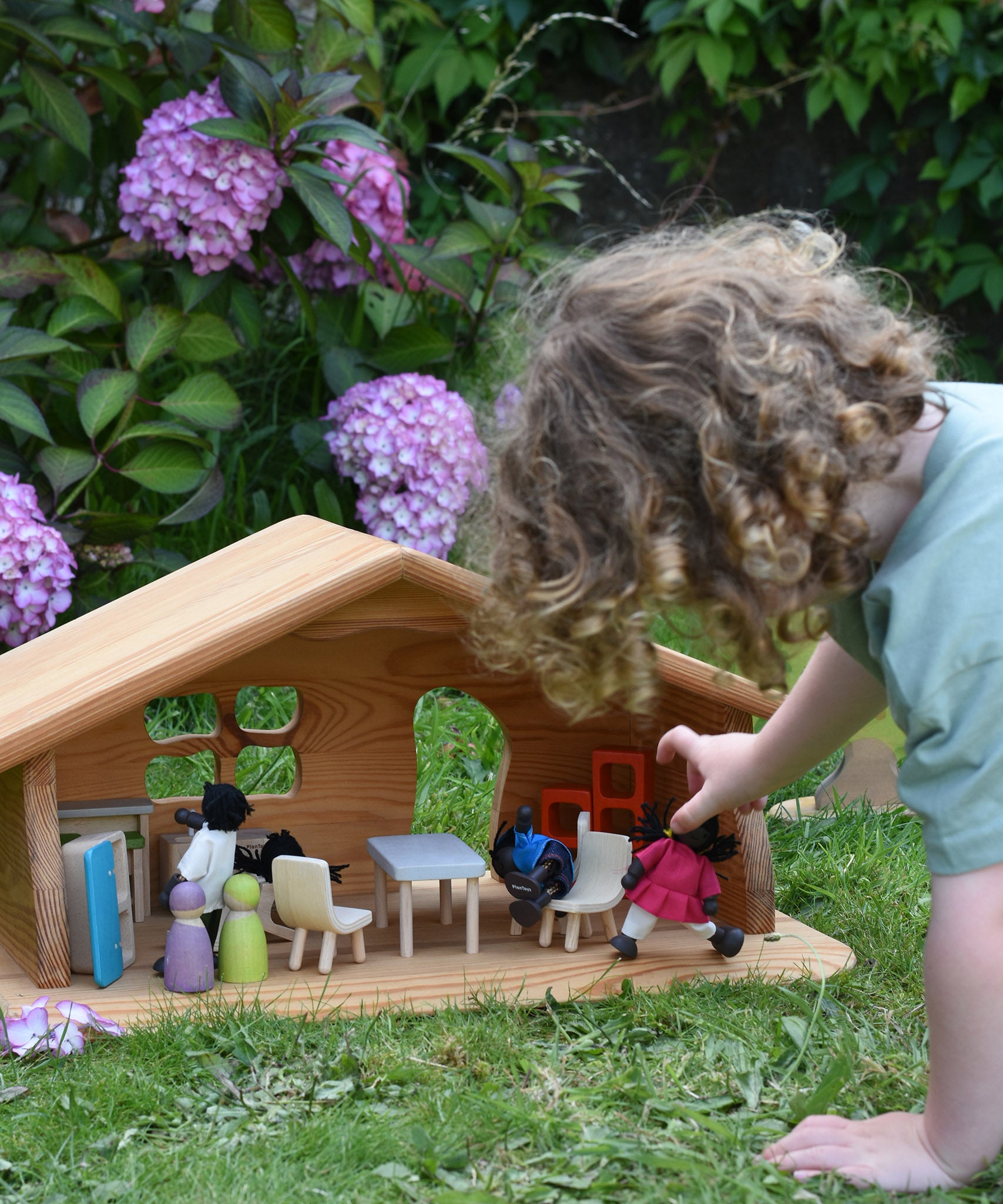 A Ostheimer wooden dolls house set up with PlanToys dolls and furniture inside. A child's hand can be seen holding one of the dolls. These wooden figures are part of a large range of expertly hand crafted, Waldorf inspired, wooden toys here at Babipur.