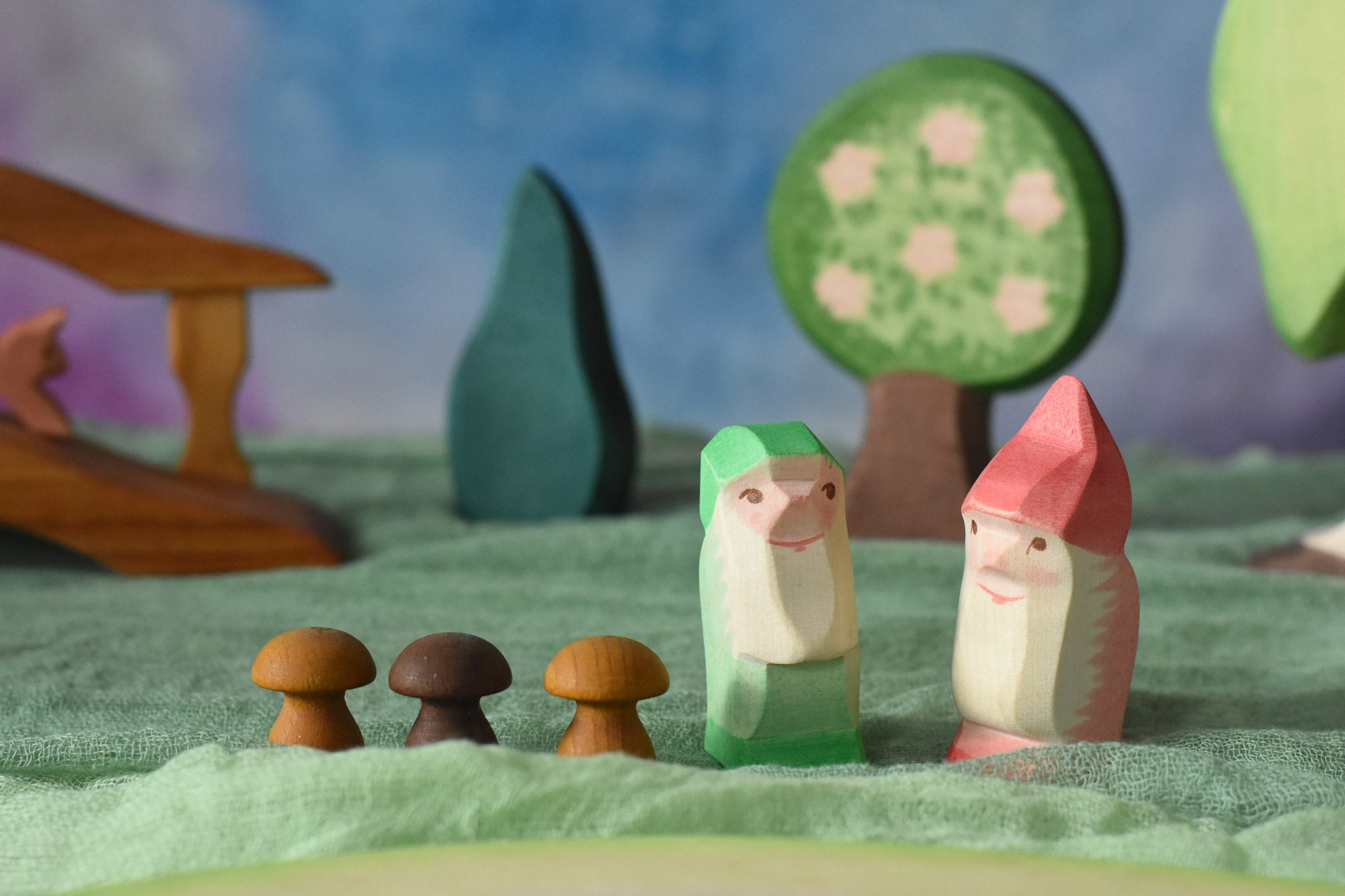 Two Ostheimer dwarves wooden toy figures placed in a small world play scene. The dwarf figures have red and green coloured outfits with matching hats and a long white beard. The figures have been placed on a green coloured Ostheimer play scarf. These wooden figures are part of a large range of expertly hand crafted, Waldorf inspired, wooden toys here at Babipur. 