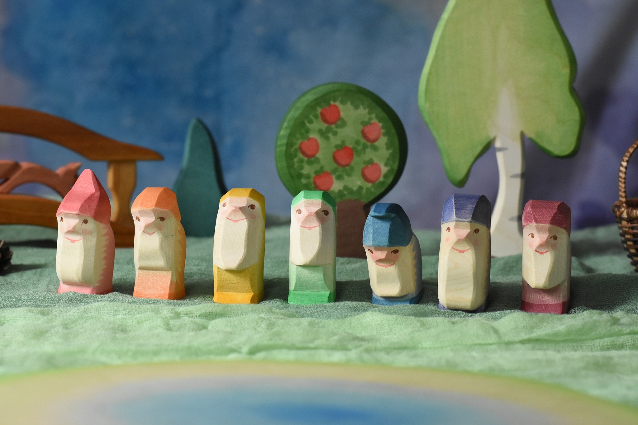 A collection of 7 coloured Ostheimer wooden dwraf wooden toy figures placed in a line in a small world play scene. There are dwarves each with a different coloured outfit, red, orange, yellow, green, blue, purple and violet. They all have matching coloured hats and white beards. These wooden figures are part of a large range of expertly hand crafted, Waldorf inspired, wooden toys here at Babipur. 