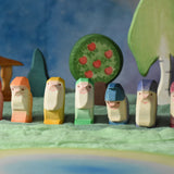 A collection of 7 coloured Ostheimer wooden dwraf wooden toy figures placed in a line in a small world play scene. There are dwarves each with a different coloured outfit, red, orange, yellow, green, blue, purple and violet. They all have matching coloured hats and white beards. These wooden figures are part of a large range of expertly hand crafted, Waldorf inspired, wooden toys here at Babipur. 