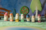 A collection of 7 coloured Ostheimer wooden dwraf wooden toy figures placed in a line in a small world play scene. There are dwarves each with a different coloured outfit, red, orange, yellow, green, blue, purple and violet. They all have matching coloured hats and white beards. These wooden figures are part of a large range of expertly hand crafted, Waldorf inspired, wooden toys here at Babipur. 