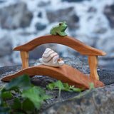 An Ostheimer Wooden sitting frog Figure placed on a railing of a wooden bridge toy  and a snail on the bridge on a stone wall outdoors. 