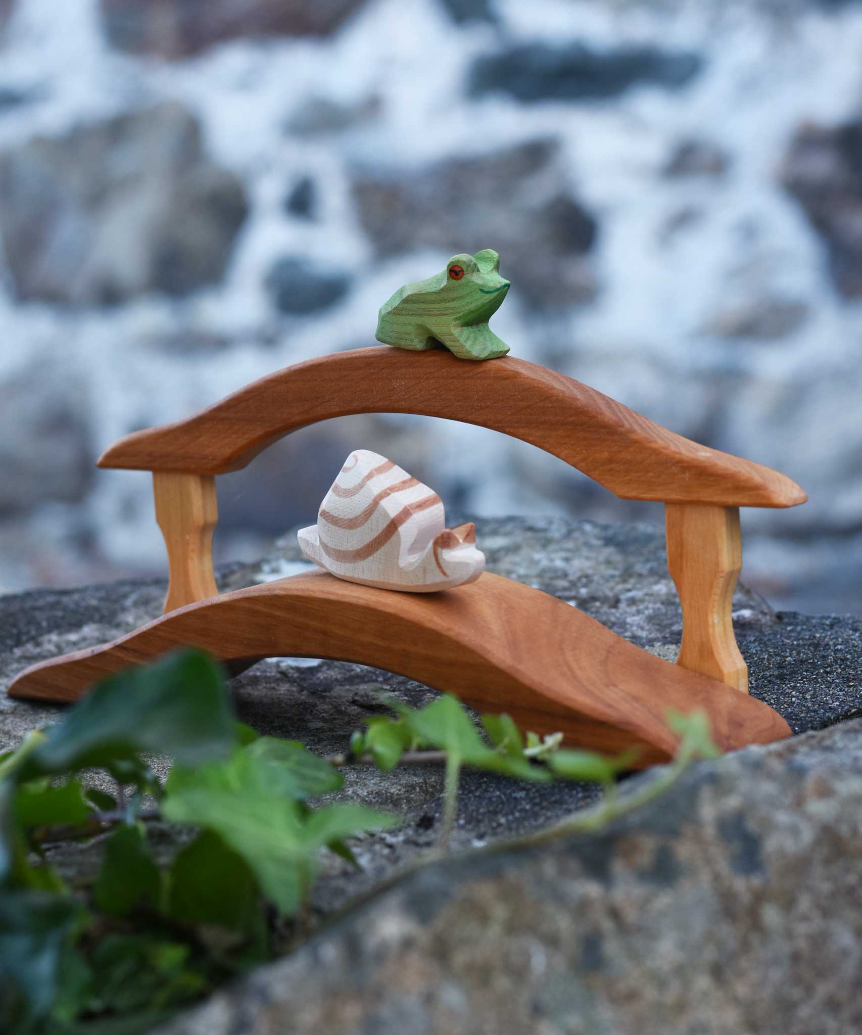 An Ostheimer Wooden sitting frog Figure placed on a railing of a wooden bridge toy  and a snail on the bridge on a stone wall outdoors. 