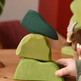 Ostheimer light, middle and dark green wooden bush scenery pieces stacked on top of each other placed on a wooden surface. Child's hand holding the bottom piece
