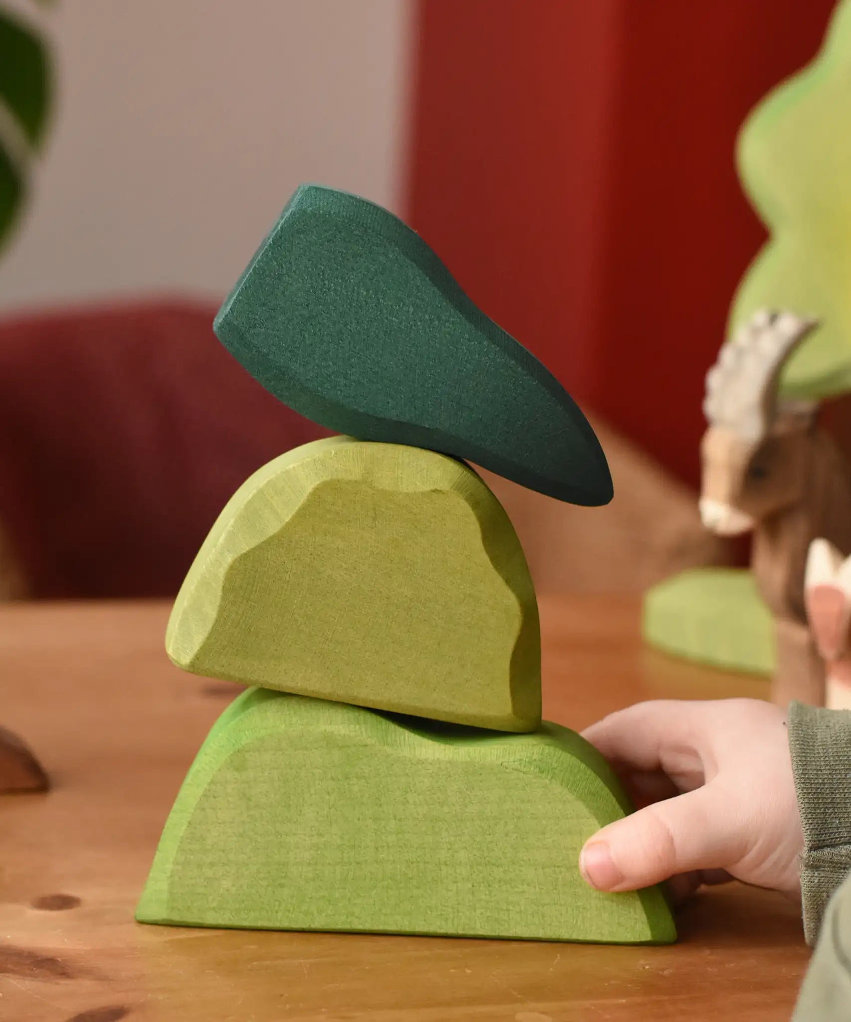 Ostheimer light, middle and dark green wooden bush scenery pieces stacked on top of each other placed on a wooden surface. Child's hand holding the bottom piece