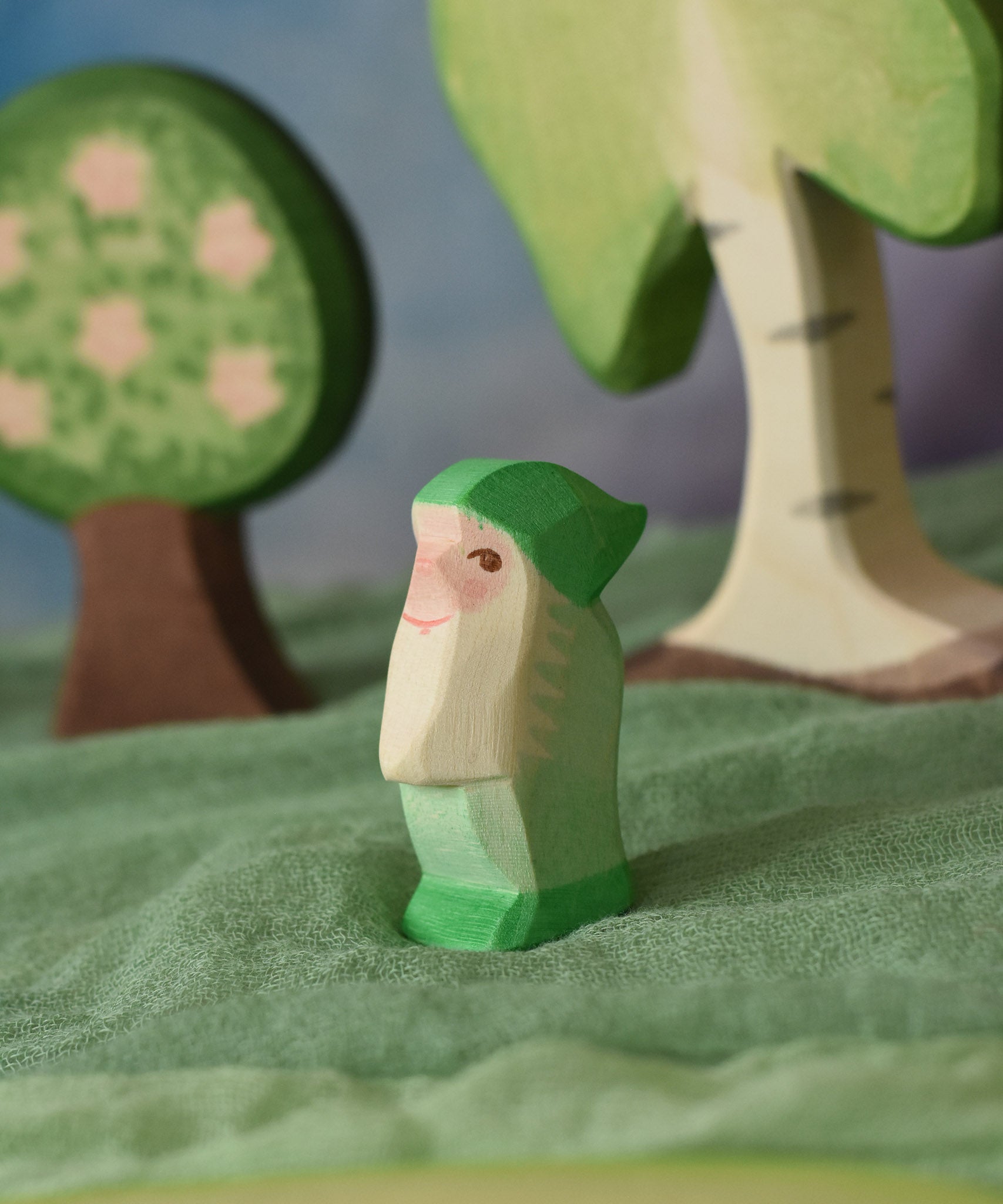 An Ostheimer green dwarf wooden toy figure placed in a small world play scene. The dwarf figure has a green coloured outfit with matching hat and a long white beard. The figure has been placed on a green coloured Ostheimer play scarf. This wooden figure is part of a large range of expertly hand crafted, Waldorf inspired, wooden toys here at Babipur. 