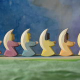 A side profile view of a host of little Ostheimer angels placed in a line on an Ostheimer pond piece. These little angel figures all have different coloured dresses, white, yellow, orange, purple, pink and blue with moon shaped wings.