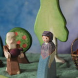 An Ostheimer wooden toy market woman figure placed in a small world scene set up. The figure has a blue coloured dress with a purple shawl and head covering. There is an Ostheimer beggar in the background. This wooden figure is part of a large range of expertly hand crafted, Waldorf inspired, wooden toys here at Babipur. 