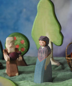 An Ostheimer wooden toy market woman figure placed in a small world scene set up. The figure has a blue coloured dress with a purple shawl and head covering. There is an Ostheimer beggar in the background. This wooden figure is part of a large range of expertly hand crafted, Waldorf inspired, wooden toys here at Babipur. 