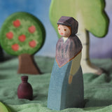 An Ostheimer wooden toy market woman figure placed in a small world scene set up. The figure has a blue coloured dress with a purple shawl and head covering. This wooden figure is part of a large range of expertly hand crafted, Waldorf inspired, wooden toys here at Babipur. 