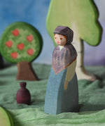 An Ostheimer wooden toy market woman figure placed in a small world scene set up. The figure has a blue coloured dress with a purple shawl and head covering. This wooden figure is part of a large range of expertly hand crafted, Waldorf inspired, wooden toys here at Babipur. 