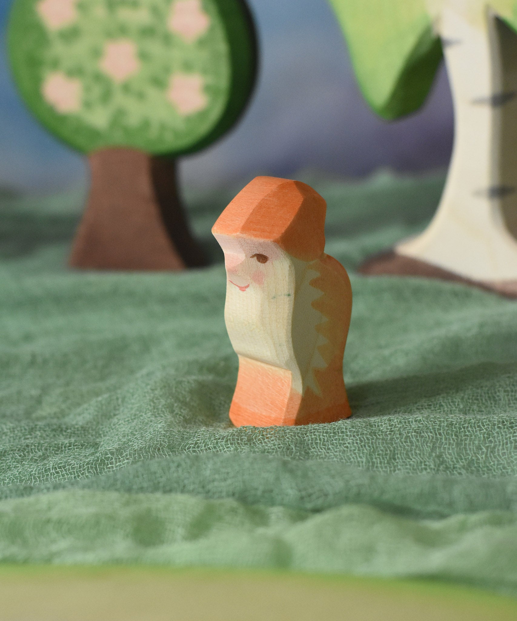 An Ostheimer orange dwarf wooden toy figure placed in a small world play scene. The dwarf figure has an orange coloured outfit with matching hat and a long white beard. The figure has been placed on a green coloured Ostheimer play scarf. This wooden figure is part of a large range of expertly hand crafted, Waldorf inspired, wooden toys here at Babipur. 