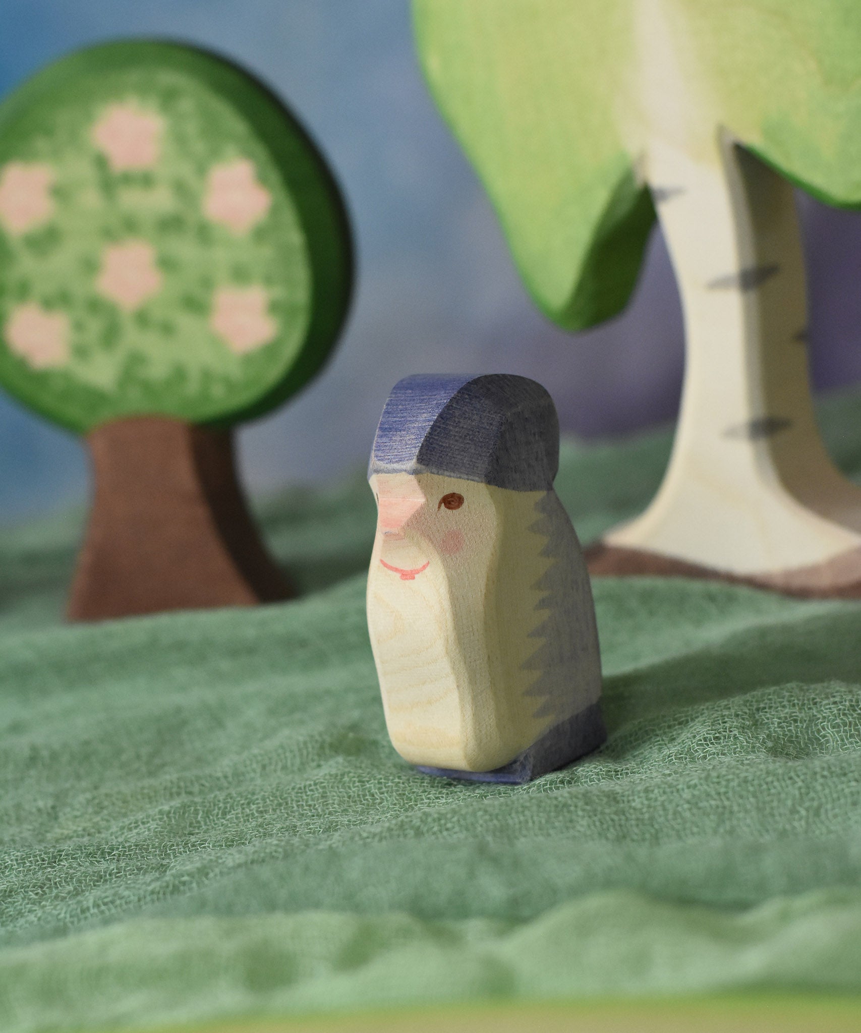 An Ostheimer purple dwarf wooden toy figure placed in a small world play scene. The dwarf figure has a purple coloured outfit with matching hat and a long white beard. The figure has been placed on a green coloured Ostheimer play scarf. These wooden figures are part of a large range of expertly hand crafted, Waldorf inspired, wooden toys here at Babipur. 