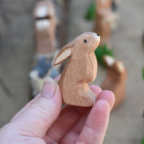 An Ostheimer wooden toy rabbit figure with it's ears low pictured in an adult's hand to show scale. Check out our full range of expertly crafted wooden toy figures and scenery here at Babipur.