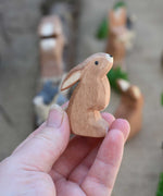 An Ostheimer wooden toy rabbit figure with it's ears low pictured in an adult's hand to show scale. Check out our full range of expertly crafted wooden toy figures and scenery here at Babipur.