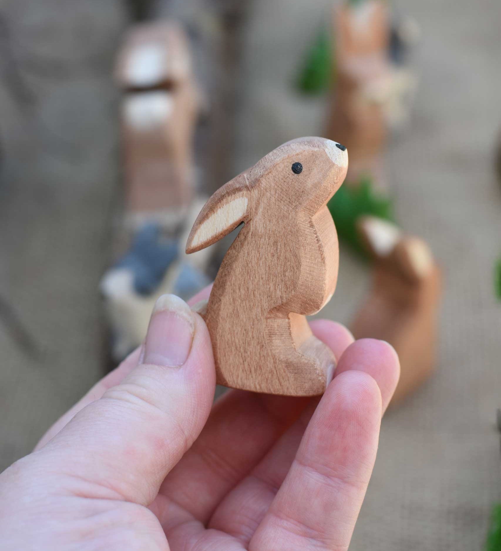 An Ostheimer wooden toy rabbit figure with it's ears low pictured in an adult's hand to show scale. Check out our full range of expertly crafted wooden toy figures and scenery here at Babipur.