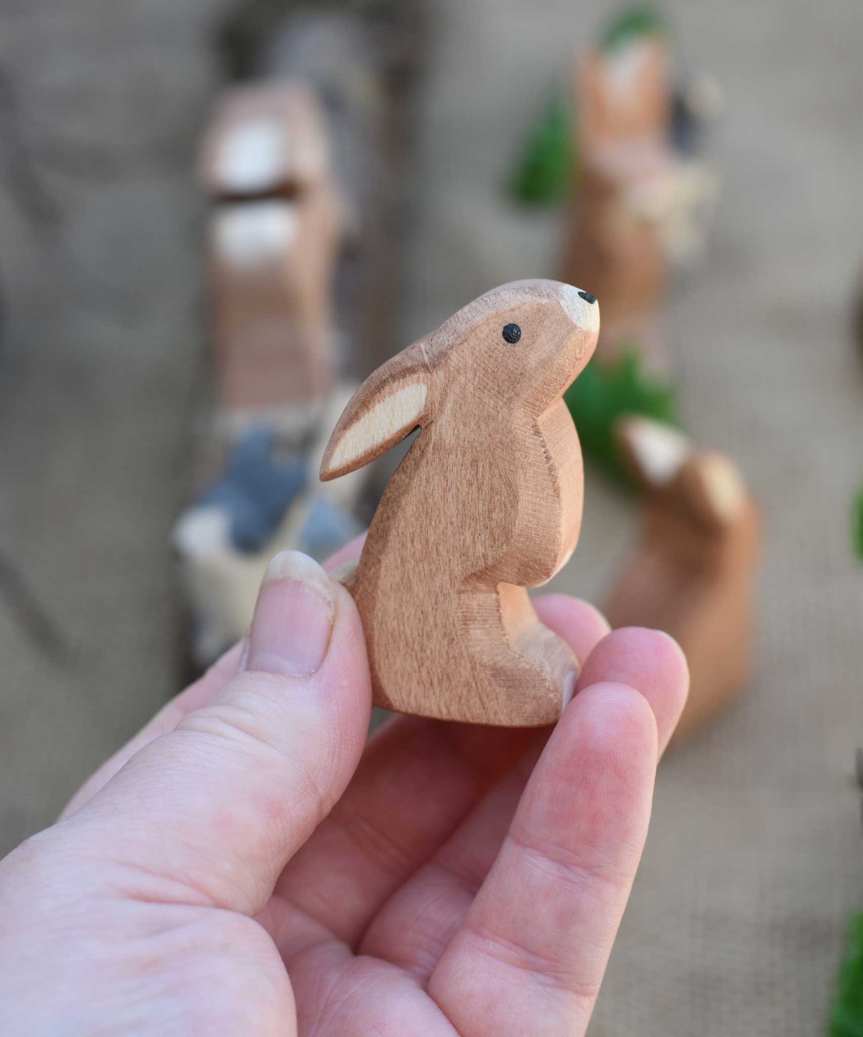 An Ostheimer wooden toy rabbit figure with it's ears low pictured in an adult's hand to show scale. Check out our full range of expertly crafted wooden toy figures and scenery here at Babipur.
