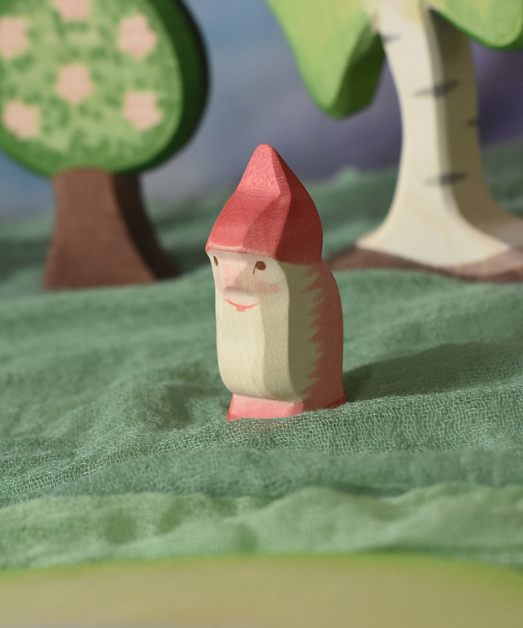 An Ostheimer red dwarf wooden toy figure placed in a small world play scene. The dwarf figure has a red coloured outfit with matching hat and a long white beard. The figure has been placed on a green coloured Ostheimer play scarf. This wooden figure is part of a large range of expertly hand crafted, Waldorf inspired, wooden toys here at Babipur. 
