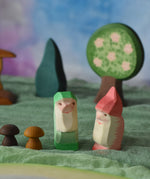 Two Ostheimer dwarves wooden toy figures placed in a small world play scene. The dwarf figures have red and green coloured outfits with matching hats and a long white beard. The figures have been placed on a green coloured Ostheimer play scarf. These wooden figures are part of a large range of expertly hand crafted, Waldorf inspired, wooden toys here at Babipur. 