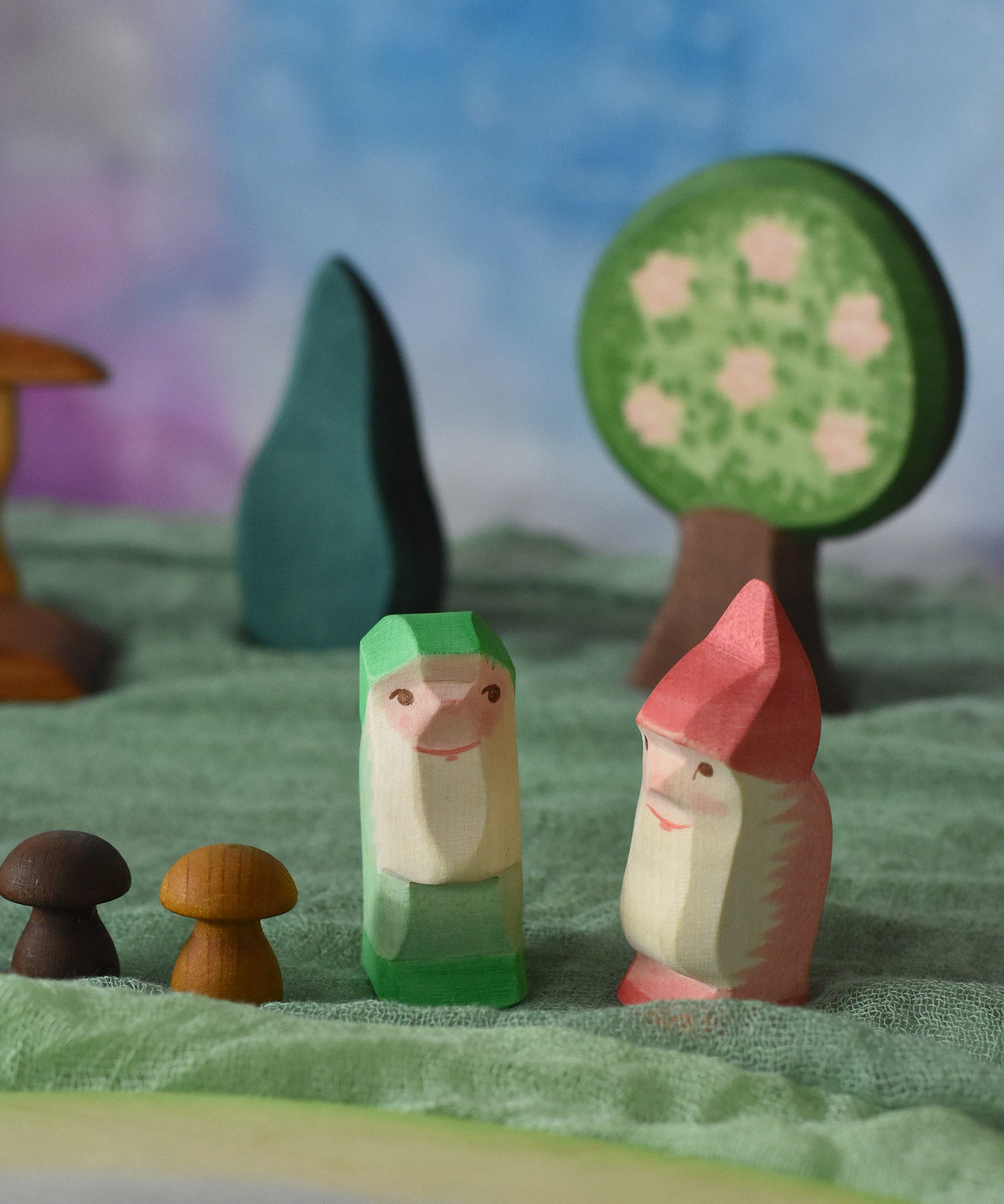 Two Ostheimer dwarves wooden toy figures placed in a small world play scene. The dwarf figures have red and green coloured outfits with matching hats and a long white beard. The figures have been placed on a green coloured Ostheimer play scarf. These wooden figures are part of a large range of expertly hand crafted, Waldorf inspired, wooden toys here at Babipur. 