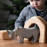 The large Ostheimer rhino wooden toy figure placed on a wooden surface. A child can be seen looking on excitedly in the background. These wooden figures are part of a wide range of expertly hand crafted wooden toys here at Babipur.