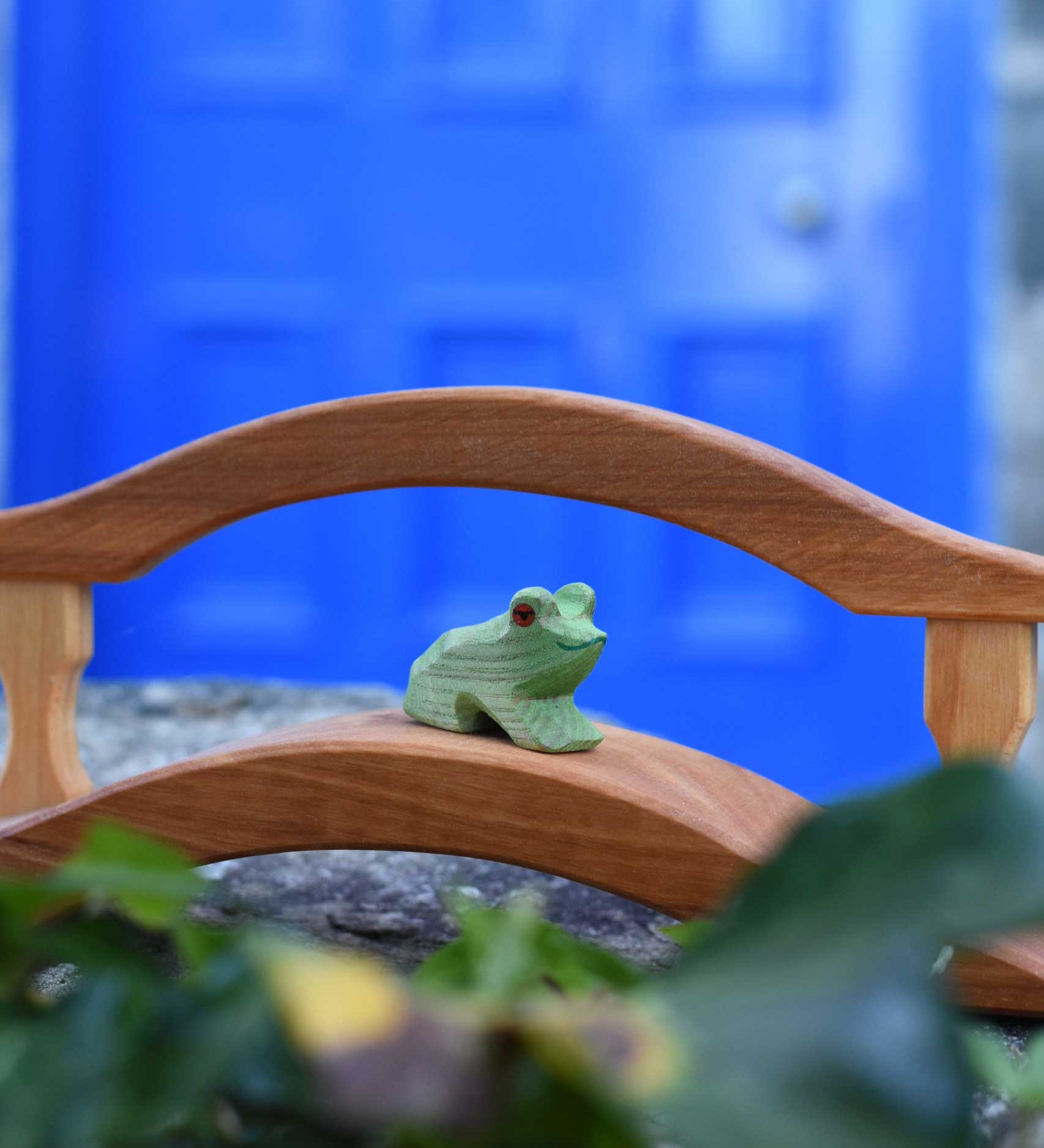 An Ostheimer Wooden sitting frog Figure placed on a wooden bridge toy on a stone wall outdoors. 