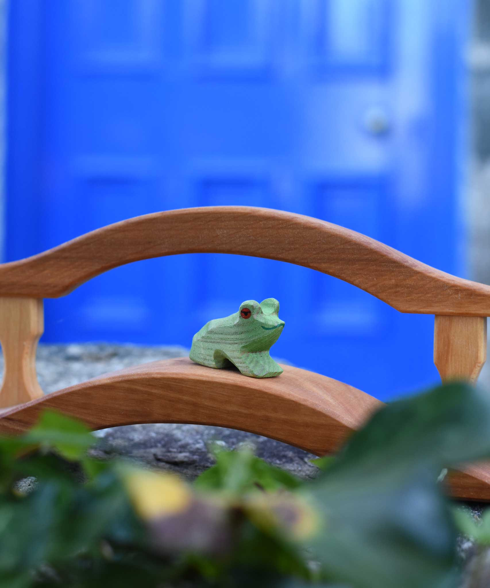An Ostheimer Wooden sitting frog Figure placed on a wooden bridge toy on a stone wall outdoors. 