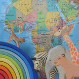 A collection of Ostheimer wooden animal figures placed next to a Grimm's rainbow in front of a world map. There is a monkey, zebra, elephant, lioness and giraffe.