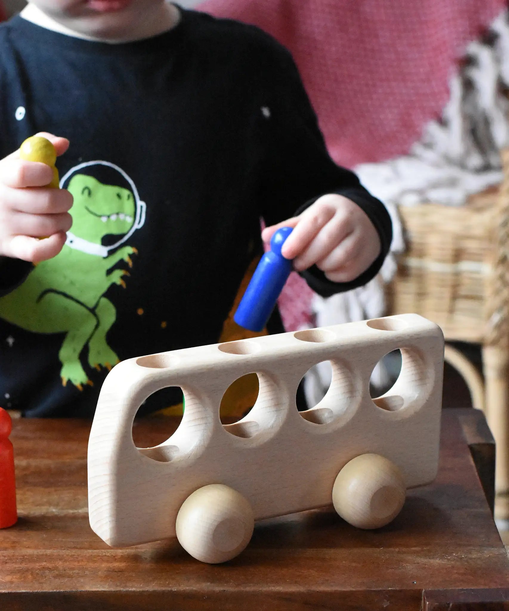 Close up of a child playing with the Ostheimer Natural Bus With 4 People. The child is placing the blue coloured wooden people figure into one of the slots at the top of the bus. 
