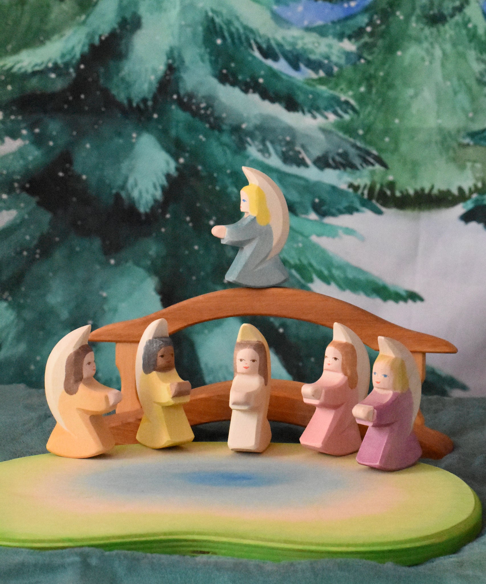 A festive scene featuring Ostheimer little angels in 6 different colours placed around the Ostheimer pond scene. The Ostheimer wooden bridge with railing can be seen in the background with a Wonderie WonderCloth with the Christmas tree design hung up on the wall behind. These wooden figures are part of a wide range of expertly hand crafted wooden toys available here at Babipur. 